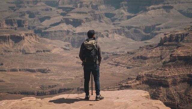 Standing hiker overlooking vast layered canyon from sandstone ledge with backpack and pole