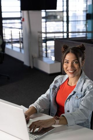 Smiling young woman typing on laptop in modern coworking office wearing denim jacket