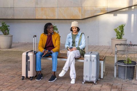 Young diverse travelers waiting on bench with rolling luggage checking phone in courtyard