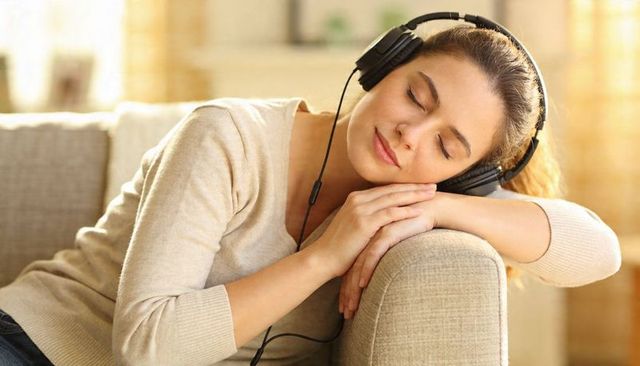 Young woman relaxing on beige sofa wearing over-ear headphones, listening and resting