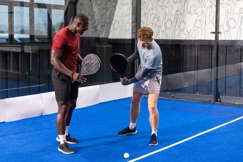 Two male friends practicing padel on vibrant blue indoor court, coaching and teamwork
