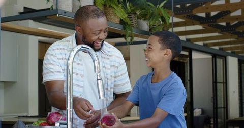 African american father and son washing onions at kitchen sink, smiling and bonding