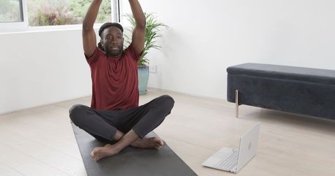 African American man practicing yoga at home following laptop in minimalist living room