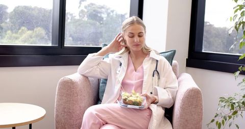 Female clinician taking lunch break in staff lounge wearing pink scrubs and white coat