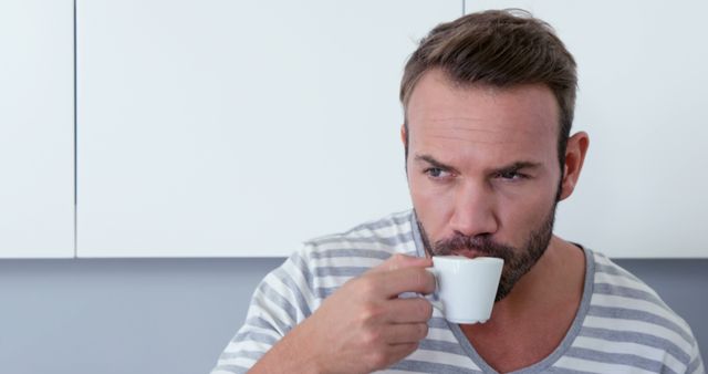 Middle-aged man drinking espresso in a modern kitchen. He is wearing a casual striped shirt and appears thoughtful. This can be used for concepts like morning routines, coffee enjoyment, relaxation at home, or solo time. It suits lifestyle and domestic settings with modern, minimalist themes.