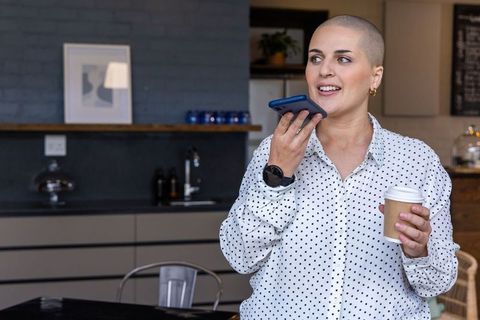 Bald woman in polka dot blouse speaking into phone while holding coffee in modern cafe kitchen