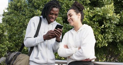 Multicultural students checking smartphone on campus railing, smiling in casual hoodies