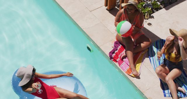 Group of friends enjoying a sunny day by the poolside. One person is floating on a pool float with a drink in hand, while two others are sitting on towels, playing with a beach ball and enjoying beverages. This image can be used for content related to summer activities, poolside relaxation, vacation promotions, and social gatherings.