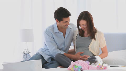 A couple is sitting on their bed, joyfully bonding over an expected arrival, surrounded by a cozy home setting. This video can be used for promotions on family planning, prenatal care, home and lifestyle editorial, and parenting blogs.