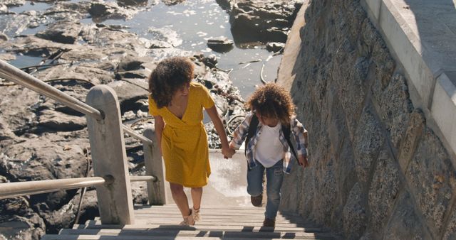 Happy biracial mother and son holding hands walking up steps from beach in the sun, copy space. Motherhood, childhood, togetherness, summer, vacations and free time, unaltered.