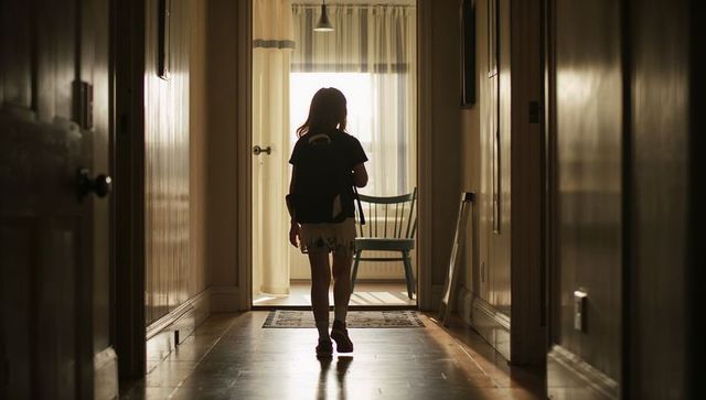 Young girl walking down home hallway toward sunlit room with backpack and wooden chair
