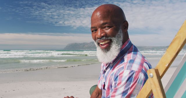 Smiling senior man with a white beard relaxing at the beach in a plaid shirt. Ideal for themes related to retirement, relaxation, vacation, leisure, healthy lifestyles, and enjoying nature.