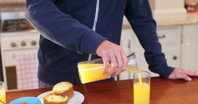 This image shows a person pouring fresh orange juice from a carafe into a glass in a kitchen. Perfect for use in articles or ads related to healthy living, breakfast ideas, home dining, casual mornings, or kitchen products. The presence of muffins complements themes of cozy and nourishing routines.