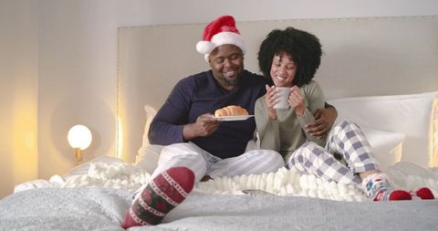 Couple sharing breakfast in bed wearing plaid pajamas, santa hat and festive holiday socks