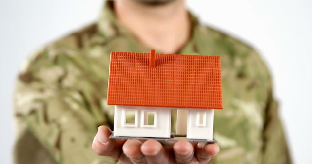 A person in military uniform holds a small model house in their hands, with copy space. Highlighting the concept of home ownership or housing security for military personnel, the image evokes themes of stability and protection.