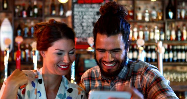 Friends smiling while watching a video on a smartphone in a bar environment full of drinks. Ideal for usage in stories about social life, weekend activities, technology in social settings or bar events, emphasizing togetherness and joy.