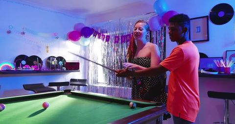 Diverse couple playing pool at neon birthday party guiding cue technique in game room