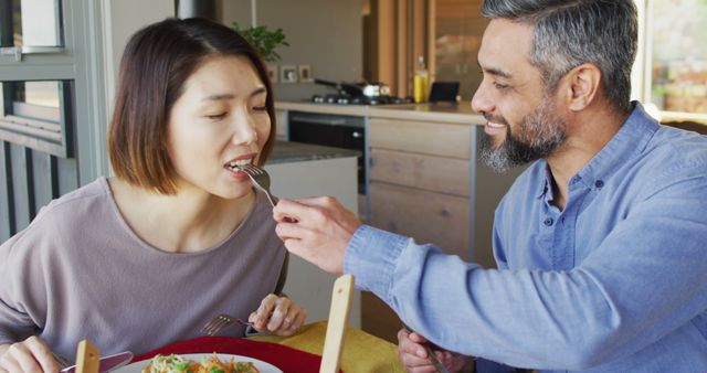 Happy couple bonding over lunch in a modern kitchen, with a middle-aged man feeding a smiling asian woman. Great for relationship advice articles, family bonding themes, or illustrating home lifestyle content.