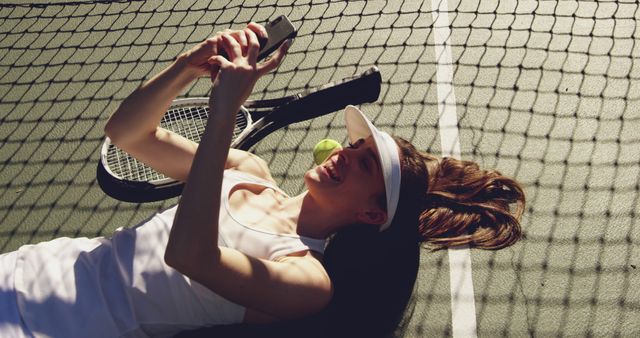 Young female tennis player lying on tennis court surface checking her smartphone. Dressed in white outfit with sun visor, tennis racquet, and ball next to her. Ideal for concepts related to fitness, sports, relaxation, leisure activities, and modern technology use.