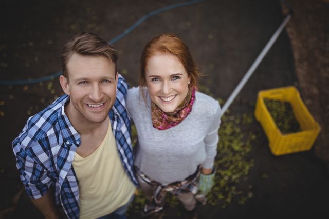 High angle view of a happy young couple standing at an olive farm, smiling at the camera. Ideal for use in agricultural promotions, rural lifestyle blogs, and advertisements focusing on sustainable living and teamwork in farming.