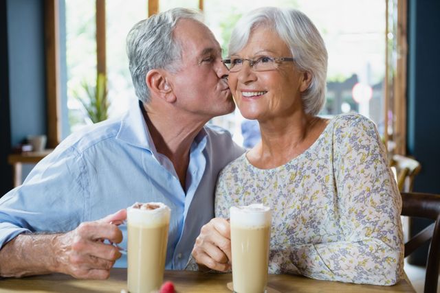Senior couple enjoying a coffee date in a cozy café. The man is kissing the woman on the cheek, both are smiling and holding lattes. Perfect for use in advertisements for retirement communities, lifestyle blogs, or promotions for cafés and coffee shops.