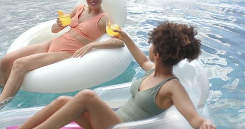 Two women relaxing on inflatable floats clinking drinks and floating in sunny pool paradise