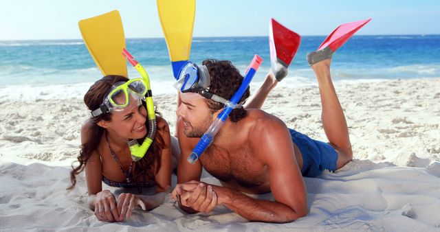 Couple lying on sandy beach, wearing snorkel gear with blue ocean in background. Perfect for travel brochures, summer vacation advertisements, tropical destination promotions, and leisure activity guides.
