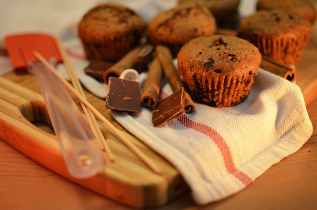 This image depicts freshly baked chocolate muffins surrounded by cinnamon sticks and chocolate pieces on a wooden cutting board. Ideal for food blogs, baking tutorials, recipe books, culinary websites, and advertisements for bakeries. The warm tone gives it a cozy, homely vibe.