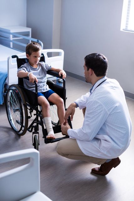 Doctor kneeling and talking to young child in wheelchair in hospital ward. Suitable for healthcare, medical, pediatric care, and patient support themes. Can be used in articles, brochures, and websites related to medical care, child health, and hospital services.