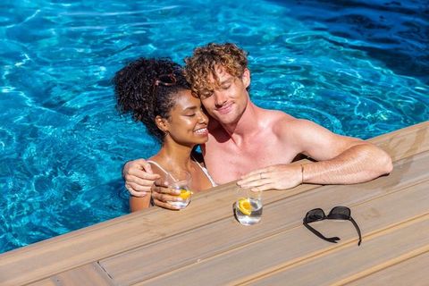 Diverse couple relaxing and embracing at pool edge holding citrus drinks on sunny deck