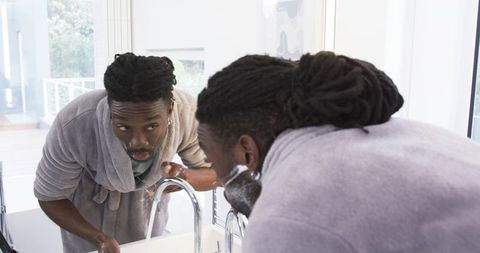 African American man shaving in bright modern bathroom wearing bathrobe at sink