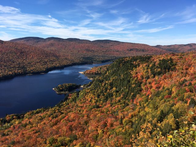 The image captures a breathtaking autumn landscape featuring vibrant fall foliage surrounding a serene lake situated between picturesque mountains. Clear blue skies and colorful trees make this an idyllic natural scene. Ideal for use in travel brochures, nature blogs, or as backgrounds in presentations, emphasizing themes of tranquility, outdoor adventure, and seasonal beauty.