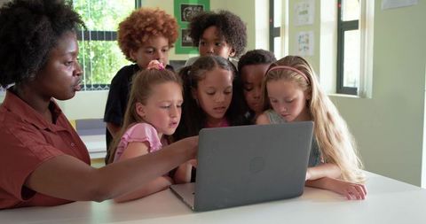Female teacher guiding diverse elementary students around laptop in bright classroom