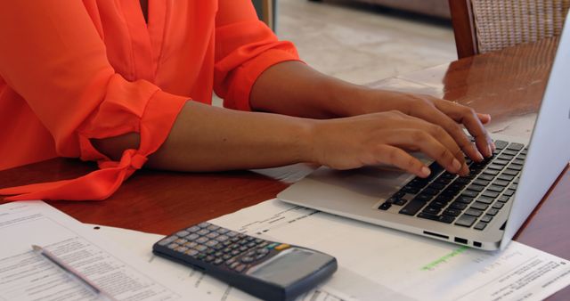 Person wearing orange sleeves typing on laptop with documents and calculator on desk. The image conveys productivity, finance management, and office work. Useful for illustrating freelance work, studying, accounting, business planning, telecommuting, or online learning. Ideal for websites, blogs, or advertisements related to work, productivity, and technology.
