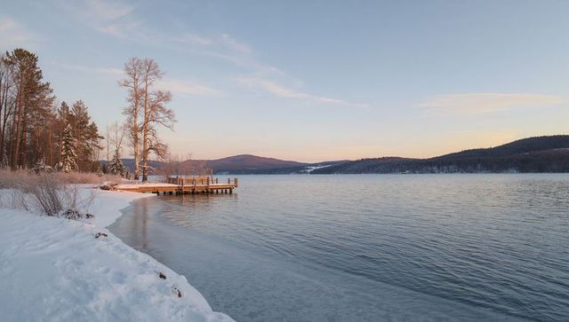 Sunrise Wooden Dock Extending into Calm Frozen Lake with Snowy Shore and Pine Trees at Dawn
