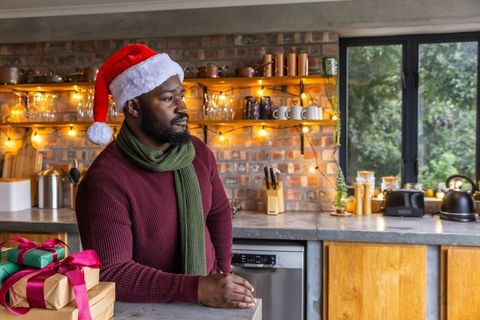 Adult african american man wearing santa hat leaning on kitchen countertop holding gifts