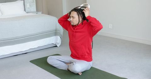 African american man sitting cross-legged on yoga mat putting on headphones in red hoodie
