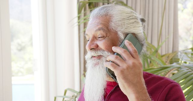 Happy biracial senior man with white beard on smartphone in sunny room, copy space, slow motion. Communication, retirement, domestic life and senior lifestyle, unaltered.