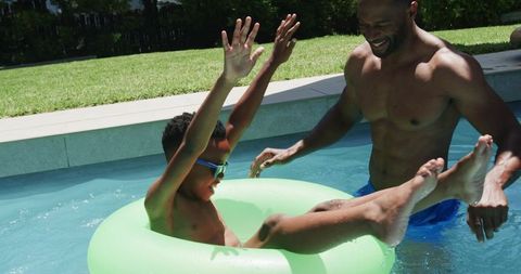 African father pushing excited son on green inflatable ring in backyard pool during summer