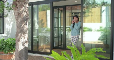 Morning calm asian woman standing at sliding glass door holding coffee in plaid pajamas