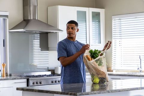 Young man placing reusable mesh grocery bag on stone island in bright modern kitchen for meal prep