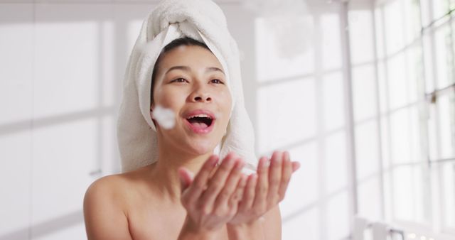 Image of portrait of smiling biracial woman with towel on hair blowing foam in bathroom. Health and beauty, leisure time, domestic life and lifestyle concept.