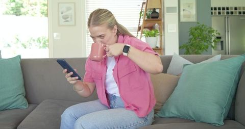 Young woman sipping coffee while checking smartphone on cozy living room sofa wearing smartwatch