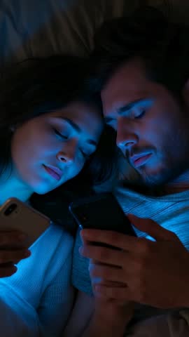 Couple scrolling smartphones in bed under blue screen glow sharing late-night intimacy