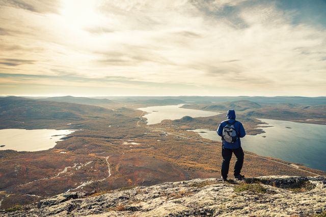 Hiker standing on mountain peak enjoying stunning sunrise view over expansive landscape with lakes below. Ideal for topics on outdoor adventure, nature exploration, solo travel, inspiring journeys, and recreational activities promotion.