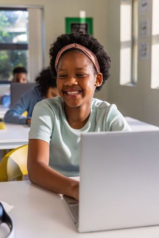 Smiling african american preteen girl typing on laptop in bright classroom for remote learning