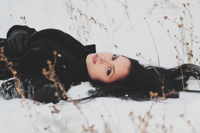 A relaxed woman is lying in the snow, wearing a black winter coat and gloves. Her dark hair contrasts with the white snow. This could be used for winter-themed promotions, lifestyle blogs, or seasonal greeting cards.