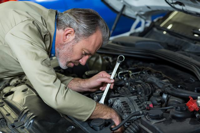 Mechanic working intently on car engine in a workshop garage, using wrench for repairs. Ideal for use in automotive service advertisements, car maintenance tutorials, professional mechanic promotions, and industrial repair content.