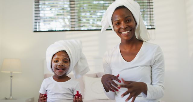 Mother and daughter with face masks and towels wrapped around their heads, smiling and spending quality time together during a skincare routine. This warm bonding moment can be used in advertisements for skincare products, articles about family activities, or promoting self-care routines.