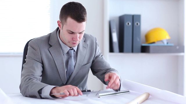 Young architect intensely focused on measuring blueprints at a tidy, organized office desk. Wearing a professional suit, he oozes concentration and determination, showcasing professionalism in the field of architecture. Background features organized files and construction safety helmet, adding to the theme of construction and planning. Perfect for business articles related to architectural planning, construction development, professional engineering scenarios, or showcasing modern office environments.